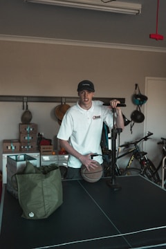 A person wearing a white shirt and a cap is standing in a room with various items. The person is holding a basketball and seems to be next to an air pump. In the background, there are bicycles and shelves with boxes. A green bag is placed on a black table, which appears to be a table tennis table.