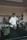 A person wearing a white shirt and a cap is standing in a room with various items. The person is holding a basketball and seems to be next to an air pump. In the background, there are bicycles and shelves with boxes. A green bag is placed on a black table, which appears to be a table tennis table.