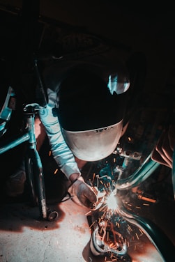 A welder in protective gear carefully shaping metal sparks flying around in a workshop.