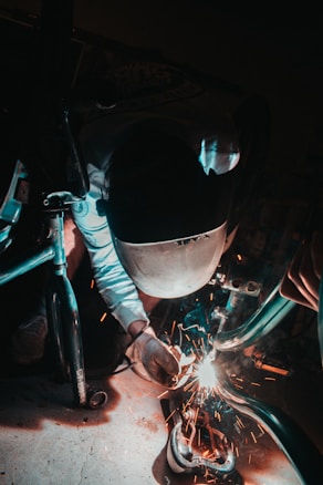 A person wearing protective gear, including a helmet and gloves, is welding metal parts with sparks flying around. The environment is dimly lit, emphasizing the bright welding sparks.