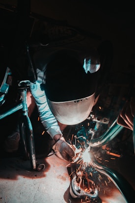 A person wearing protective gear, including a helmet and gloves, is welding metal parts with sparks flying around. The environment is dimly lit, emphasizing the bright welding sparks.