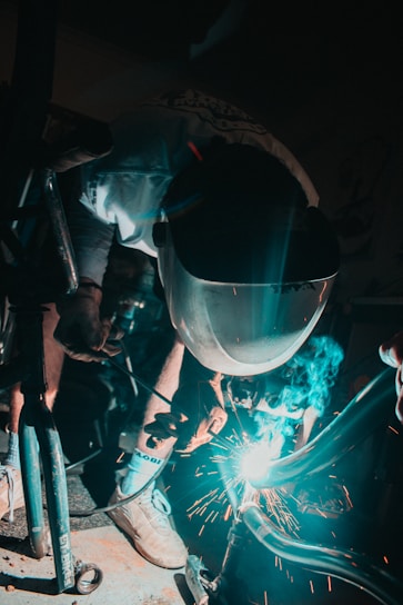 A person wearing a protective welding mask is engaged in welding metal parts, with sparks flying around. The scene is dimly lit with a focused light illuminating the welding action and casting shadows.