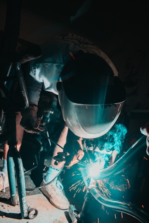 A person wearing a protective welding mask is engaged in welding metal parts, with sparks flying around. The scene is dimly lit with a focused light illuminating the welding action and casting shadows.