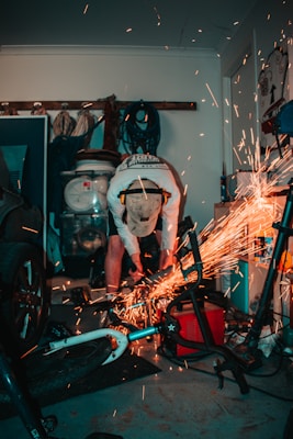 A person is working in a cluttered garage, using a tool that creates sparks as it cuts through a metal object. The surroundings include a bicycle, various tools, and equipment hanging on the wall. Protective gear like gloves and ear protection are worn by the person indicating a safety-conscious approach to the task.
