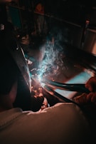A person wearing protective gear is engaged in welding. Sparks and blue light emanate from the welding process while smoke rises in the dimly lit workshop setting.