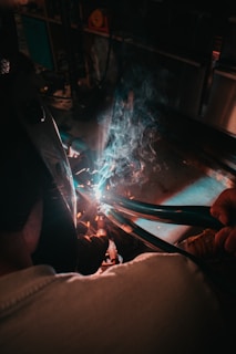 A person wearing protective gear is engaged in welding. Sparks and blue light emanate from the welding process while smoke rises in the dimly lit workshop setting.