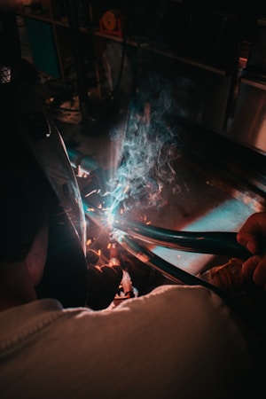 A person wearing protective gear is engaged in welding. Sparks and blue light emanate from the welding process while smoke rises in the dimly lit workshop setting.