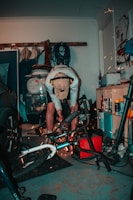 A person is working in a cluttered garage, using power tools on a bicycle. Sparks are visible as they cut or grind metal. The person is wearing a cap and ear protection. Various tools and equipment are scattered around, including a car tire, ropes, and cleaning products.
