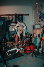 A person is working in a cluttered garage, using power tools on a bicycle. Sparks are visible as they cut or grind metal. The person is wearing a cap and ear protection. Various tools and equipment are scattered around, including a car tire, ropes, and cleaning products.