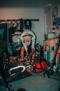 A person is working in a cluttered garage, using power tools on a bicycle. Sparks are visible as they cut or grind metal. The person is wearing a cap and ear protection. Various tools and equipment are scattered around, including a car tire, ropes, and cleaning products.