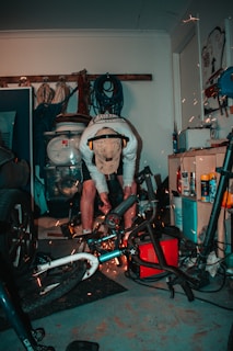 A person is working in a cluttered garage, using power tools on a bicycle. Sparks are visible as they cut or grind metal. The person is wearing a cap and ear protection. Various tools and equipment are scattered around, including a car tire, ropes, and cleaning products.