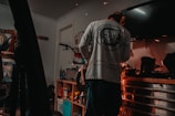 A person wearing a long-sleeve shirt and protective earmuffs stands in a dimly lit garage or workshop. The room is filled with various tools and equipment, including a bicycle mounted on the wall and a workbench cluttered with containers, spray cans, and power tools. Sparks are flying from a tool, indicating some form of metalwork or repair is being done.