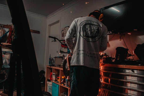 A person wearing a long-sleeve shirt and protective earmuffs stands in a dimly lit garage or workshop. The room is filled with various tools and equipment, including a bicycle mounted on the wall and a workbench cluttered with containers, spray cans, and power tools. Sparks are flying from a tool, indicating some form of metalwork or repair is being done.