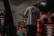 A person wearing a long-sleeve shirt and protective earmuffs stands in a dimly lit garage or workshop. The room is filled with various tools and equipment, including a bicycle mounted on the wall and a workbench cluttered with containers, spray cans, and power tools. Sparks are flying from a tool, indicating some form of metalwork or repair is being done.