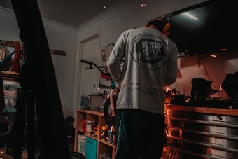 A person wearing a long-sleeve shirt and protective earmuffs stands in a dimly lit garage or workshop. The room is filled with various tools and equipment, including a bicycle mounted on the wall and a workbench cluttered with containers, spray cans, and power tools. Sparks are flying from a tool, indicating some form of metalwork or repair is being done.