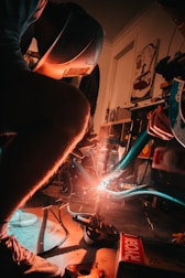 A person wearing a welding helmet is engaged in welding metal, possibly part of a bicycle frame. Sparks are flying around the area, and the scene is dimly lit with a warm orange glow from the welding. The setting appears to be a workshop with various tools and equipment visible in the background.