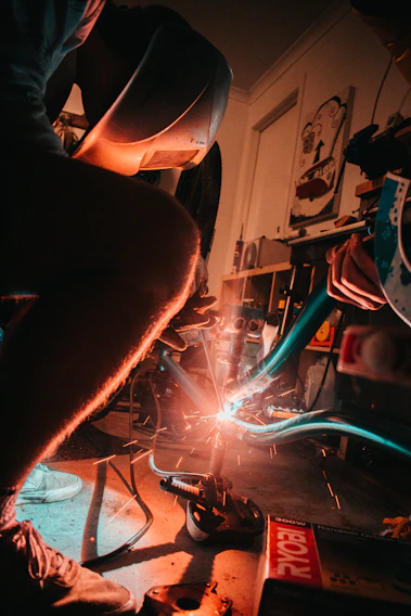 Close-up of a craftsman welding a sleek black matte steel frame with sparks flying in a dimly lit workshop.