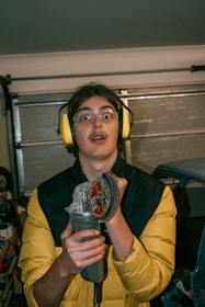 A person wearing protective yellow earmuffs and clear safety glasses is holding an angle grinder. The background appears to be a garage with a partially open garage door.