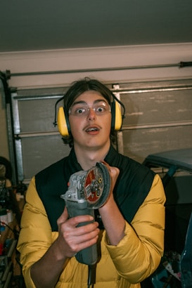A person wearing protective yellow earmuffs and clear safety glasses is holding an angle grinder. The background appears to be a garage with a partially open garage door.