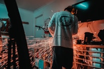 A person is wearing protective ear gear while engaging in metal grinding, creating bright sparks that illuminate the workspace. The setting appears to be a garage or workshop with various tools and equipment scattered around. The lighting is dim, adding a dramatic effect to the scene.