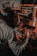 A person wearing a welding mask is engaged in a workshop environment, working with metal pieces. Sparks fly as the person works, suggesting some form of welding or metal cutting activity. The workspace is cluttered with tools and materials, including a toolbox and a tire visible in the background.