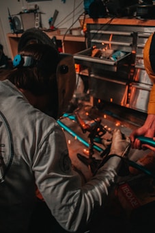 A person wearing a welding mask is engaged in a workshop environment, working with metal pieces. Sparks fly as the person works, suggesting some form of welding or metal cutting activity. The workspace is cluttered with tools and materials, including a toolbox and a tire visible in the background.