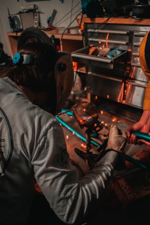 A person wearing a welding mask is engaged in a workshop environment, working with metal pieces. Sparks fly as the person works, suggesting some form of welding or metal cutting activity. The workspace is cluttered with tools and materials, including a toolbox and a tire visible in the background.