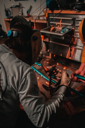 A person wearing a welding mask is engaged in a workshop environment, working with metal pieces. Sparks fly as the person works, suggesting some form of welding or metal cutting activity. The workspace is cluttered with tools and materials, including a toolbox and a tire visible in the background.