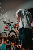 A person is working on a bicycle in a workshop environment, with visible sparks from a tool indicating metalwork or repairs being performed. Shelving behind contains various tools and products, including spray cans and a poster with a red van image.