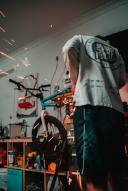 A person is working on a bicycle in a workshop environment, with visible sparks from a tool indicating metalwork or repairs being performed. Shelving behind contains various tools and products, including spray cans and a poster with a red van image.