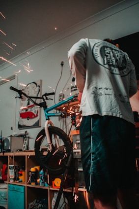 A person is working on a bicycle in a workshop environment, with visible sparks from a tool indicating metalwork or repairs being performed. Shelving behind contains various tools and products, including spray cans and a poster with a red van image.