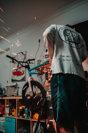 A person is working on a bicycle in a workshop environment, with visible sparks from a tool indicating metalwork or repairs being performed. Shelving behind contains various tools and products, including spray cans and a poster with a red van image.