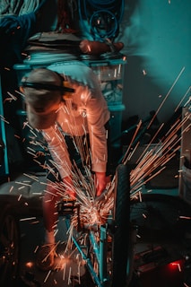 A person is focused on using a power tool, producing sparks around a bicycle in a cluttered workshop. The environment includes various tools and equipment, with bright sparks providing a stark contrast against the dimly lit setting.