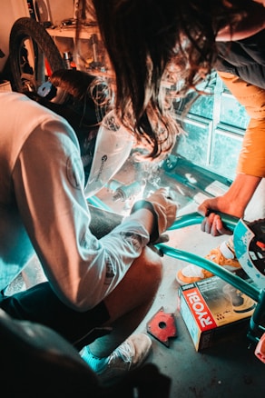Two individuals are engaged in a welding task inside a garage or workshop. One person wears a welding helmet, focusing on their work with bright sparks flying. Nearby, tools and equipment are arranged, including a box labeled 