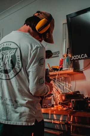 A person wearing a beige cap and earmuffs is using a power tool in a workshop. Sparks fly as the tool grinds against a metal object on a wooden workbench. The background features various tools and equipment, with a black monitor on the wall.