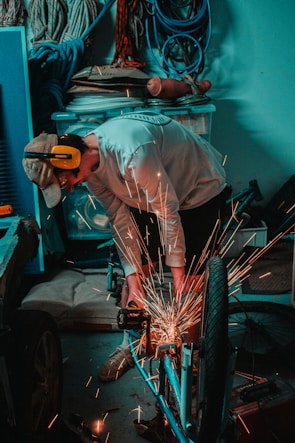 A person wearing a cap and protective ear gear is using a tool to create sparks while working on a bicycle. Various ropes, cables, and containers are stored neatly in the background, creating an organized workshop atmosphere.