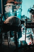 A person is bent over, working on a bicycle in a cluttered garage or workshop. Various tools and storage boxes are scattered around, and there's a stool nearby. Light casts a bluish glow on the scene, highlighting the workspace amid the disorganization.
