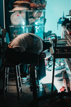 A person is bent over, working on a bicycle in a cluttered garage or workshop. Various tools and storage boxes are scattered around, and there's a stool nearby. Light casts a bluish glow on the scene, highlighting the workspace amid the disorganization.
