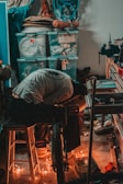 A person works in a cluttered workshop surrounded by various tools, plastic storage bins, and coiled cables. They are engaged in a task involving a bicycle, with sparks flying around, indicating some kind of metalwork or welding.