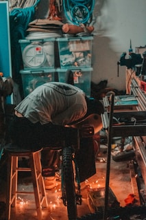 A person works in a cluttered workshop surrounded by various tools, plastic storage bins, and coiled cables. They are engaged in a task involving a bicycle, with sparks flying around, indicating some kind of metalwork or welding.