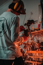 A person wearing protective gear uses a grinding tool, causing bright orange sparks to fly around a workshop space. The individual is focused on their task, wearing a long-sleeved shirt and earmuffs for safety.