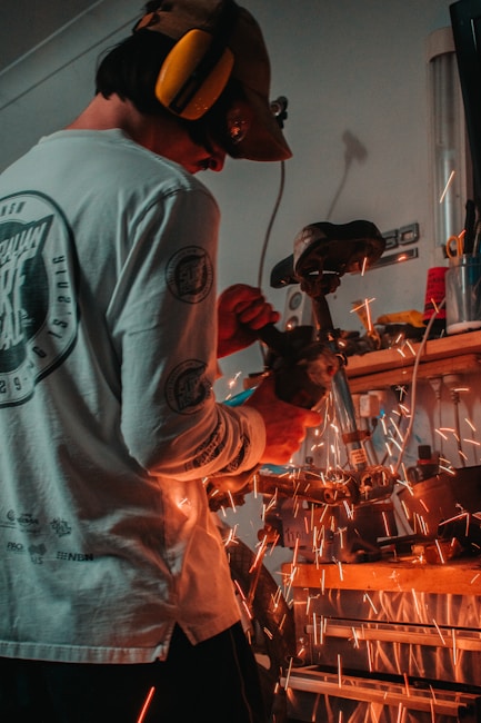 A person wearing protective gear uses a grinding tool, causing bright orange sparks to fly around a workshop space. The individual is focused on their task, wearing a long-sleeved shirt and earmuffs for safety.