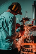 A person is working with an angle grinder in a workshop, generating bright orange sparks. The individual is wearing hearing protection and a long-sleeved shirt. Various tools and equipment are visible in the background.