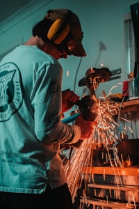 A person is working with an angle grinder in a workshop, generating bright orange sparks. The individual is wearing hearing protection and a long-sleeved shirt. Various tools and equipment are visible in the background.