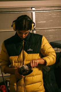A person wearing a yellow and black padded jacket and protective yellow earmuffs stands indoors, focusing on an electric power tool in their hands. The setting appears to be a garage or workshop, with a vehicle and tools in the background. The lighting is dim and warm, suggesting an industrial atmosphere.