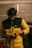 A person wearing a yellow and black padded jacket and protective yellow earmuffs stands indoors, focusing on an electric power tool in their hands. The setting appears to be a garage or workshop, with a vehicle and tools in the background. The lighting is dim and warm, suggesting an industrial atmosphere.