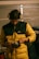 A person wearing a yellow and black padded jacket and protective yellow earmuffs stands indoors, focusing on an electric power tool in their hands. The setting appears to be a garage or workshop, with a vehicle and tools in the background. The lighting is dim and warm, suggesting an industrial atmosphere.