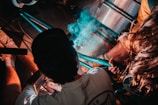 A person is engaged in welding or metalwork, with visible blue sparks and smoke emitted from the activity. The setting appears to be a workshop or garage, with metal pipes and tools surrounding the individual. Another person is assisting or observing closely. The atmosphere is dynamic with a focus on a detailed manual task.