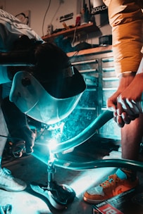 A person is engaged in welding, with sparks and light emanating from the welding tool. The room is a workshop setting with various tools and equipment visible in the background. One person's hand is steadying a metal frame, while another is wearing protective gear, including a welding mask and gloves.