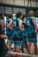 A group of young men wearing matching blue sports jerseys with tribal patterns stand indoors, likely in a gymnasium. The players are numbered, and their focus appears to be on something off-camera. A child sits on the bench nearby, adding an unexpected element to the scene.